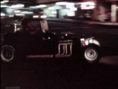 1969 Lincoln Continental - A dark-colored hot rod roadster with prominent exhaust pipes drives down a street at night.