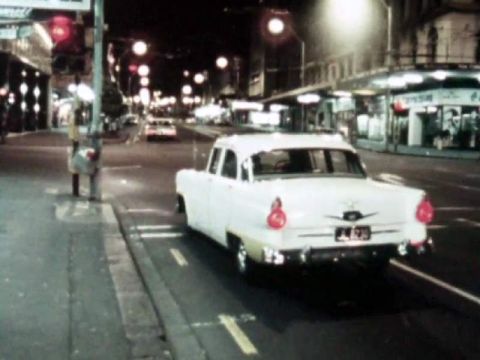 1969 Lincoln Continental - A white sedan with yellow trim drives through a city at night, with streetlights and buildings visible.
