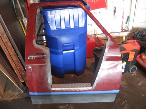 1996 Ford Bronco - A dark red truck door shell stands off the vehicle in a garage, propped against a blue trash barrel, with the window glass and inner trim r…