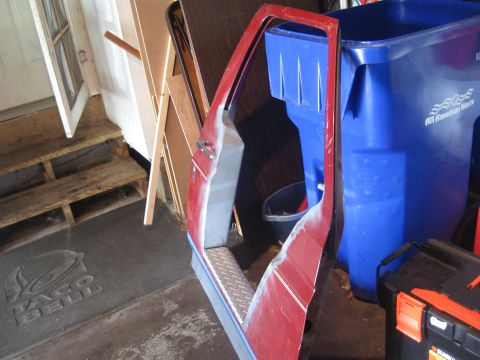 1996 Ford Bronco - A red truck door skin and cut B-pillar/rocker section propped in a home garage, showing grey primer and body filler over the repaired lower…
