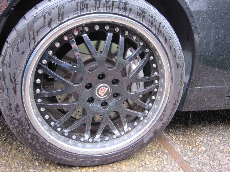 Black multi-spoke alloy wheel with chrome lip and tire on a wet surface