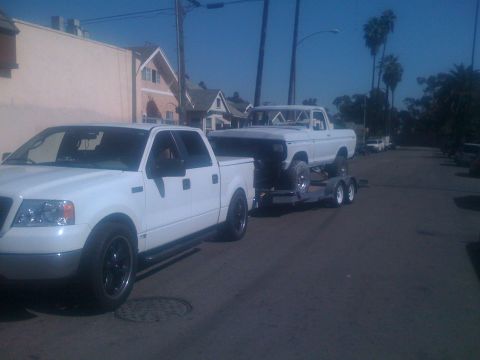 1978 Ford Bronco - A white pickup truck tows another white pickup truck on a trailer down a residential street.
