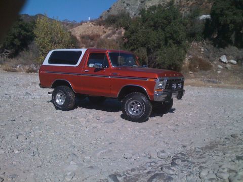 1978 Ford Bronco - This 1978 Ford Bronco, with its distinctive orange paint and white hardtop, is shown in a three-quarter front view on a gravelly terrain.