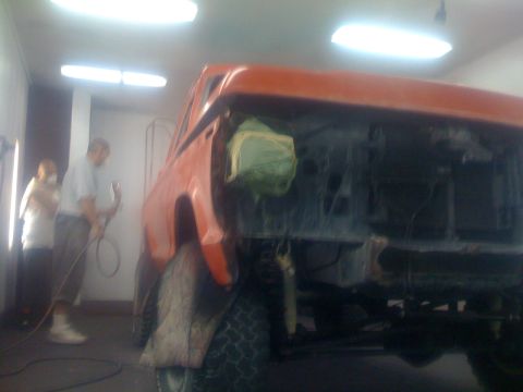 1978 Ford Bronco - An orange SUV is being worked on in a paint booth, with two people present.