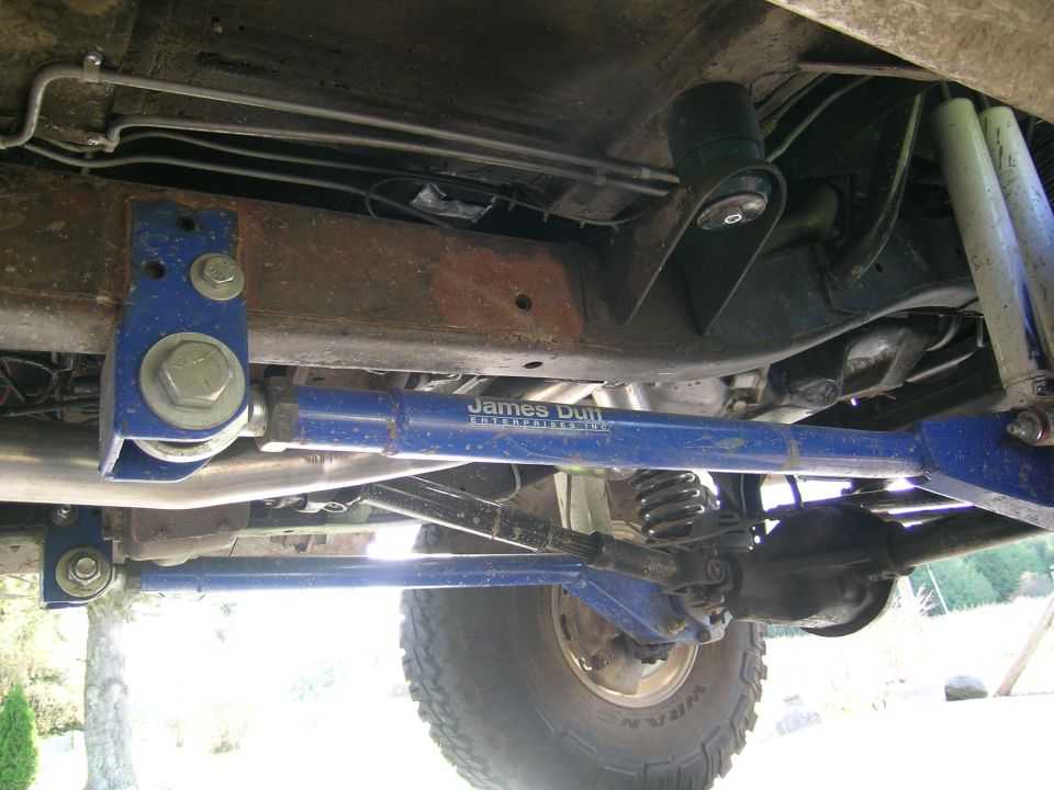 Underbody view of a vehicle showing blue aftermarket suspension arms labeled 'James Duff Enterprises' and a tire