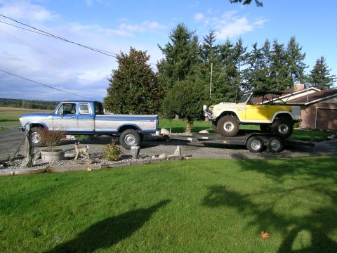 1966 Ford Bronco - A lifted Ford pickup truck is parked on blocks next to a trailer carrying a yellow Bronco under a partly cloudy sky.