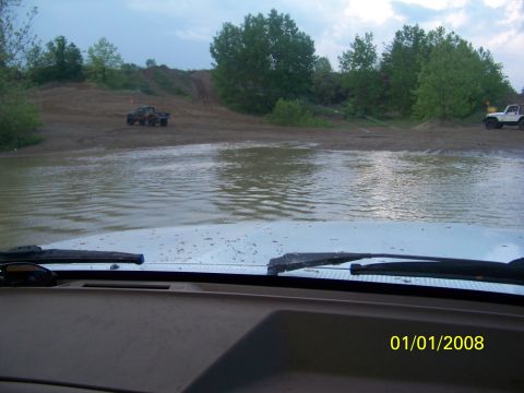 1989 Ford Bronco - Two off-road vehicles navigate a muddy water crossing on a dirt track, with trees and hills in the background.
