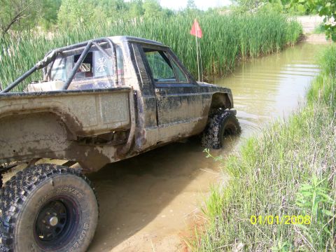 1989 Ford Bronco - A heavily modified pickup truck navigates a muddy water obstacle, with a red flag visible in the background.
