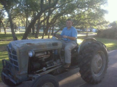 1983 Ford 427 - A man sits on a weathered tractor, which is parked outdoors on a paved surface.
