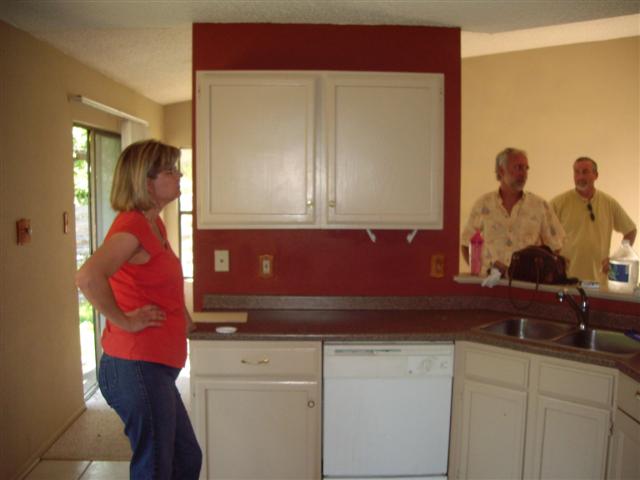 Woman in orange shirt and jeans stands in kitchen, two men behind counter