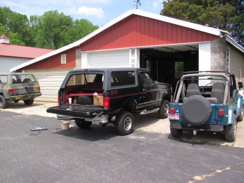 1995 Ford Bronco - Three vehicles, including a black Ford Bronco and two Jeeps, are parked outside a garage on a sunny day.