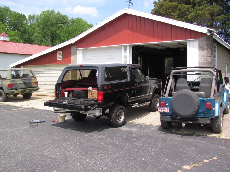 1995 Ford Bronco with tailgate open, parked next to two Jeeps by a red garage