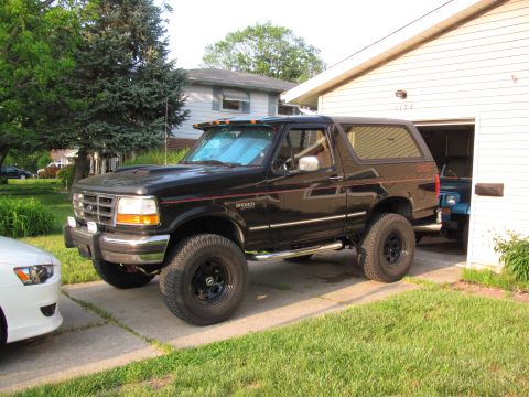 1995 Ford Bronco - 1995 Ford Bronco parked outside a garage, with a white car to its left and another vehicle visible inside the garage.