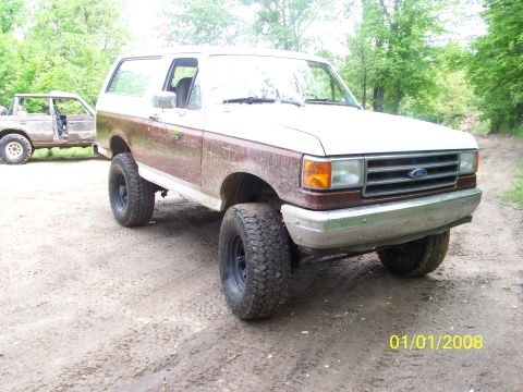 1989 Ford Bronco - A white and brown Ford Bronco is parked on a dirt road next to another vehicle.