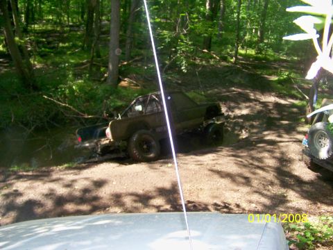 1989 Ford Bronco - Two off-road vehicles navigate a muddy trail in a wooded area.