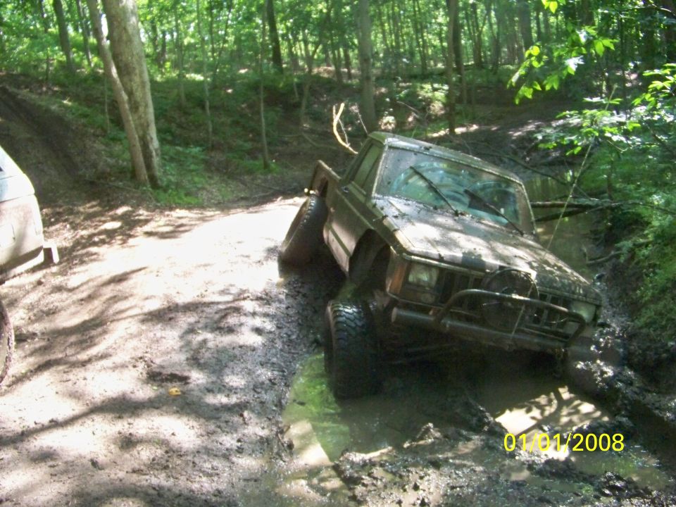 Muddy Jeep Cherokee XJ pickup truck stuck in a ditch off-road. Another vehicle is partially visible to the left