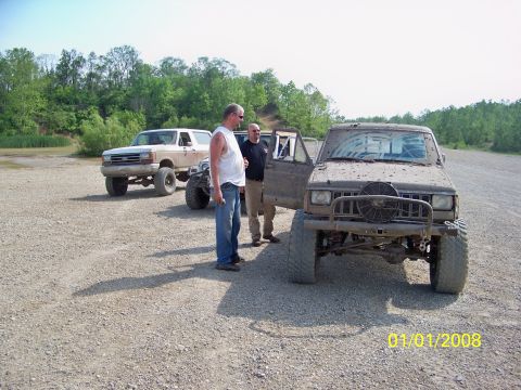 1989 Ford Bronco - Two men stand near a muddy Jeep Cherokee and a 1989 Ford Bronco on a gravel lot.