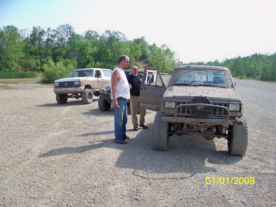 1989 Ford Bronco and a muddy Jeep Cherokee parked on gravel with two men standing between them