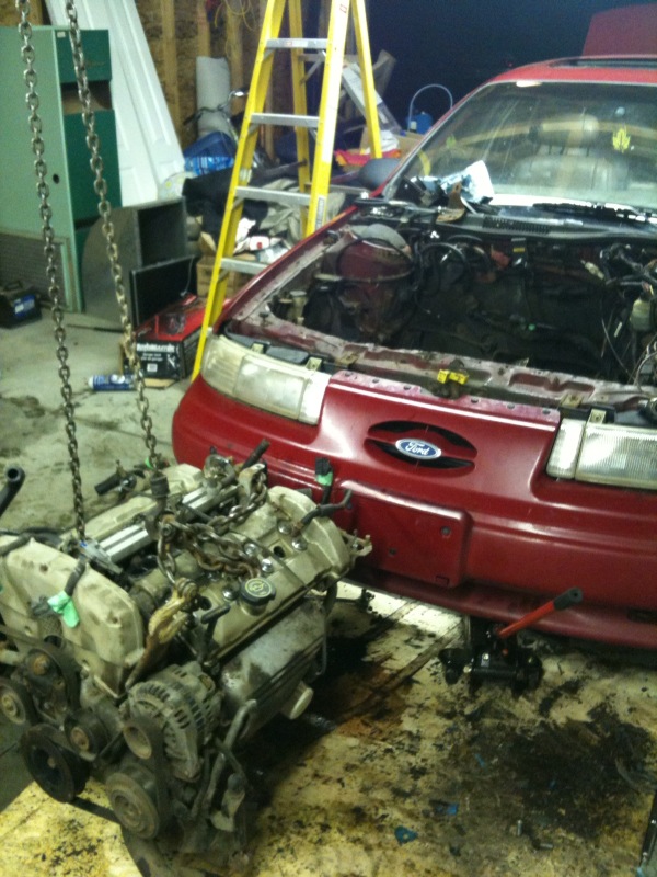 red car with hood open, engine removed and hanging from a chain hoist, ladder in background