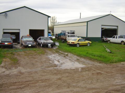 1984 Ford Mustang - Several vehicles are parked in front of industrial buildings on a cloudy day.