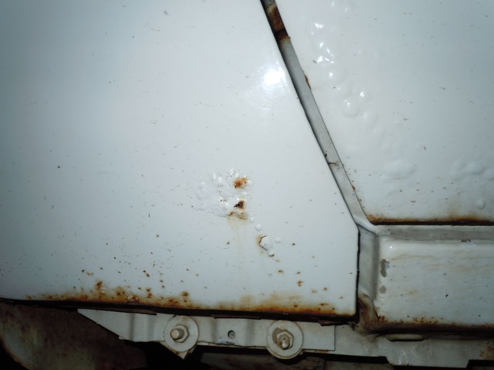 White car body panel showing rust damage along the bottom edge and two small rust spots higher up, with visible bolts and metal brackets underneath