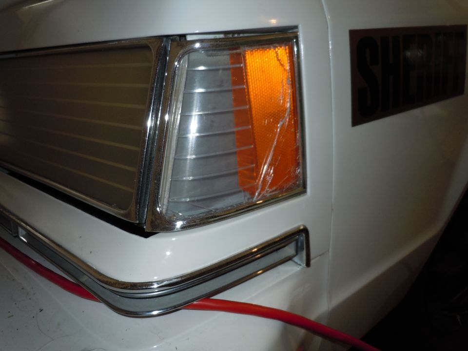 Close-up of a white police car's front corner, showing a cracked amber turn signal and the word "SHERIFF" on the side