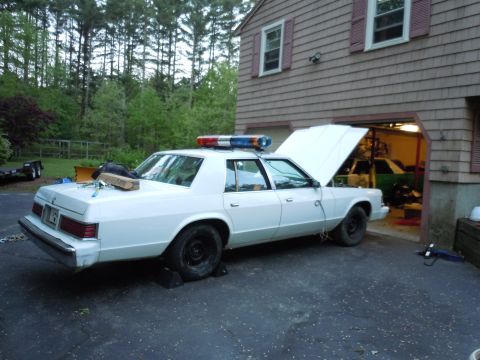 2001 Ford Crown Victoria - This older model police car, likely from the 1980s, is parked outside a garage with its hood up.