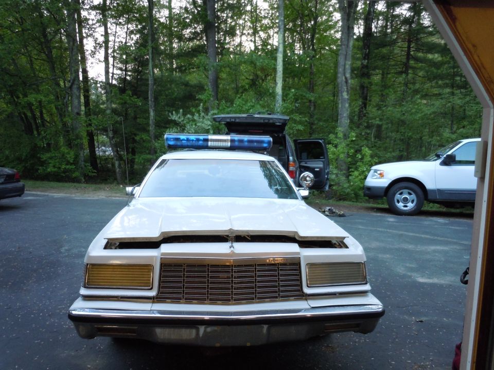 White police car with blue light bar on roof, hood open. Black SUV and white pickup truck in background