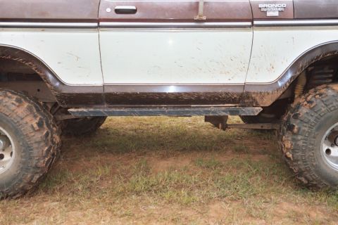1978 Ford Bronco - Side view of a 1978 Ford Bronco with rock skids and off-road tires, showing mud splatters on its brown and white exterior.