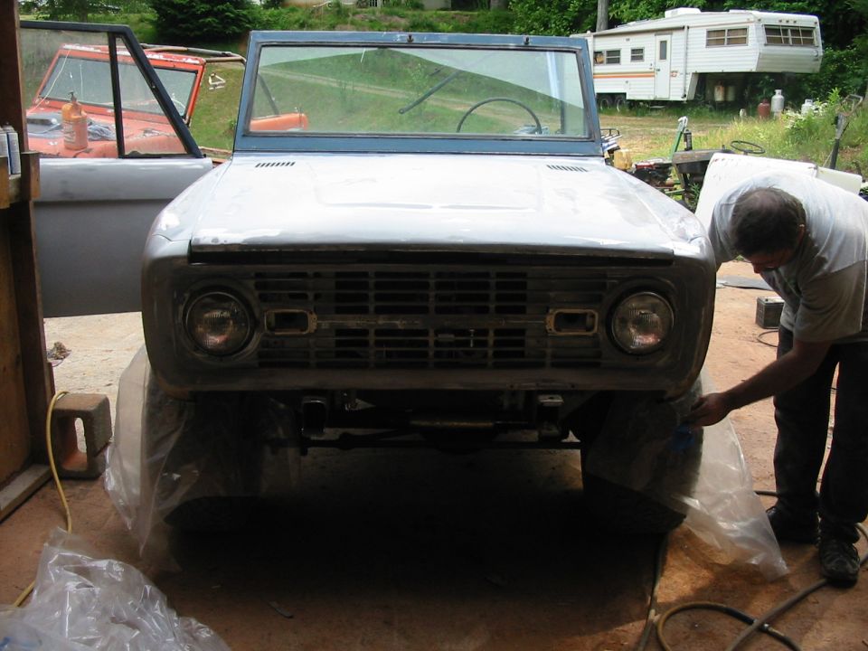 Front view of a primer-gray early Ford Bronco under restoration, man working on passenger fender