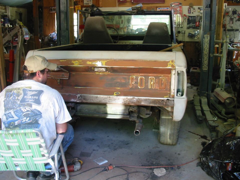 Rear of early Ford Bronco with stripped rusty tailgate showing FORD letters, man working in garage