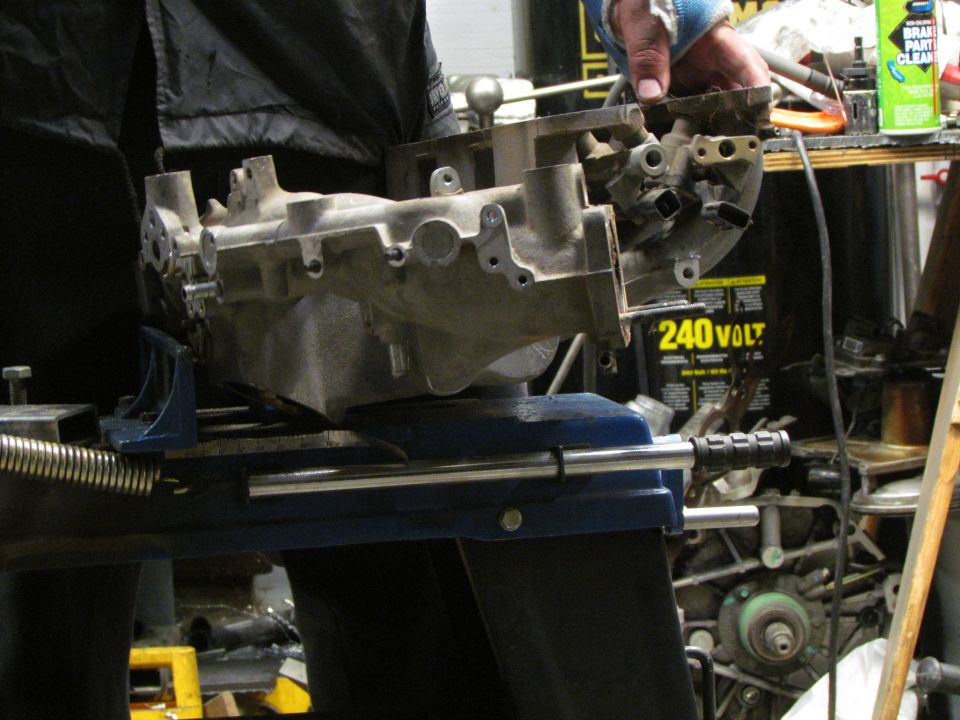 Man holding aluminum engine intake manifold clamped in a vise, workshop background