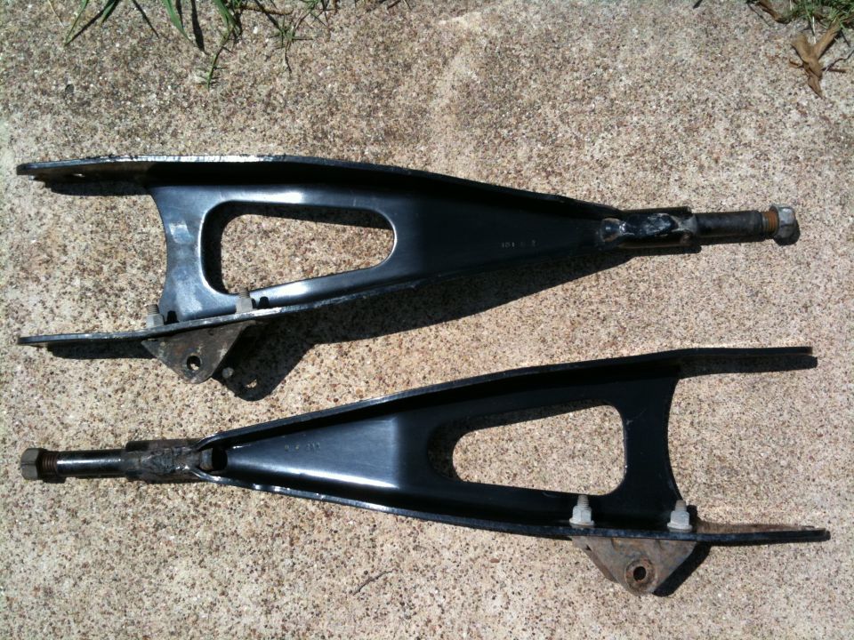 Two black control arms on concrete ground. The upper arm has a bolt on the right end. The lower arm has two white plastic clips on its bottom surface