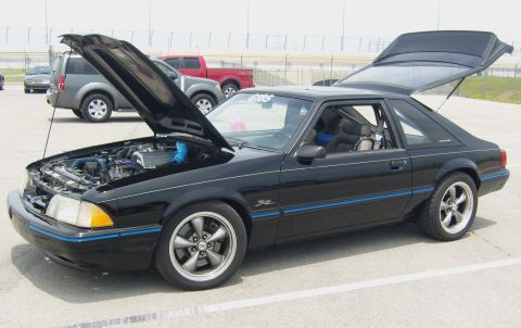 2007 Ford Mustang - A black coupe sits with its hood and trunk open, revealing the engine and interior, with other vehicles and a racetrack in the background.