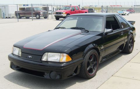2007 Ford Mustang - This black Ford Mustang is parked on asphalt, with a race track fence and other vehicles visible in the background.
