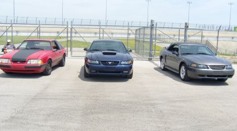 2007 Ford Mustang - Three generations of Ford Mustangs are parked side-by-side in front of a chain-link fence at a racetrack.