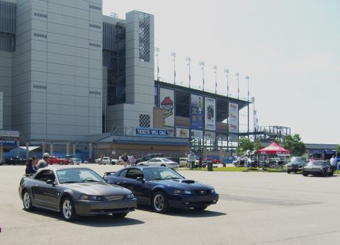 2007 Ford Mustang - Two Ford Mustangs are parked in front of a large stadium with advertisements on its side.