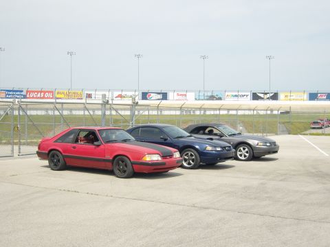 2007 Ford Mustang - Three Ford Mustangs, a red coupe and two convertibles, are parked in front of a racetrack grandstand.