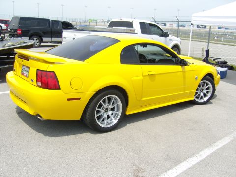 2007 Ford Mustang - A bright yellow coupe is parked on a paved surface with other vehicles and a tent in the background.