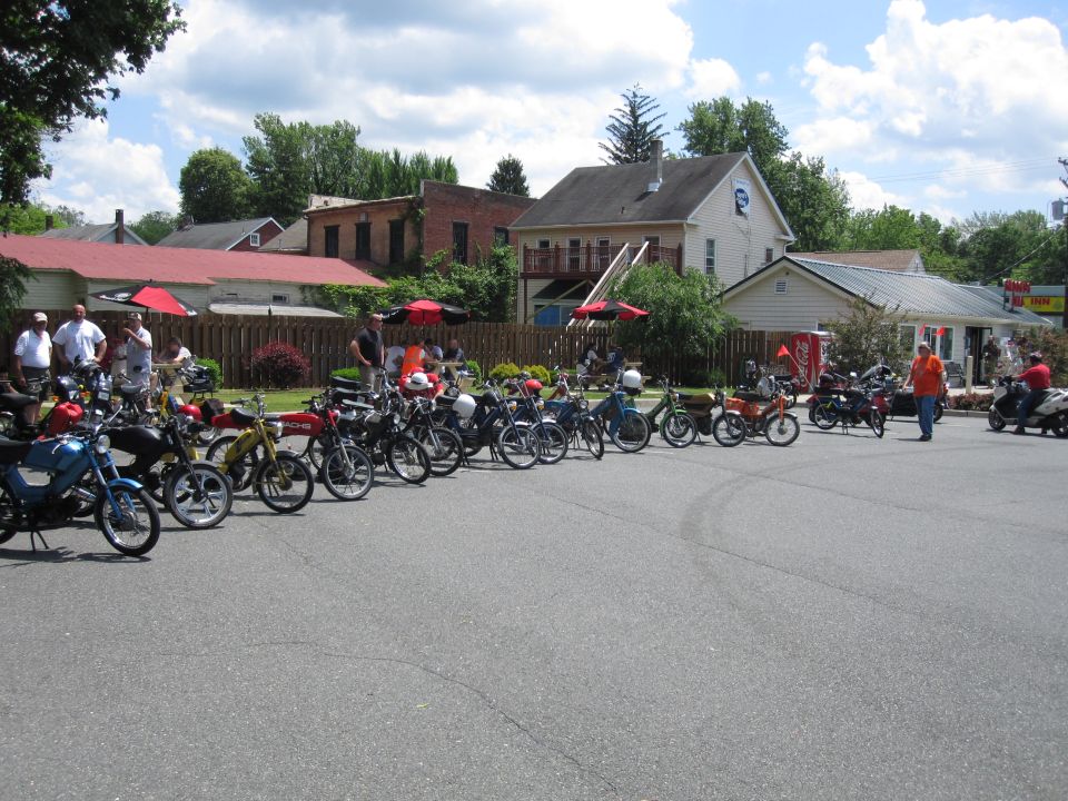 Collection of mopeds parked in a lot with buildings and trees in the background