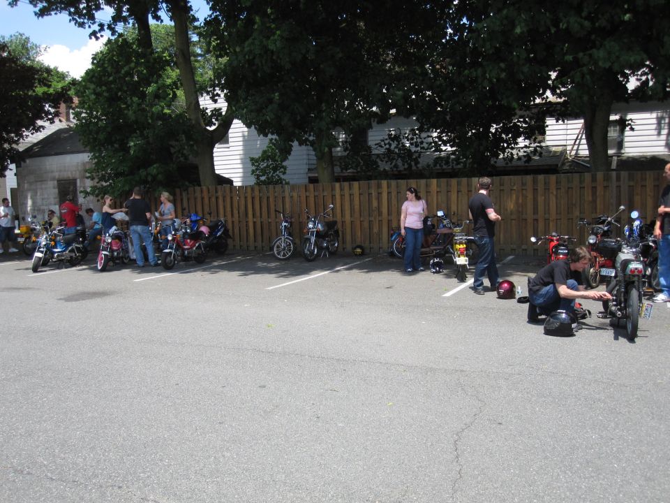 Several mopeds parked in a lot with people standing around and one person working on a moped