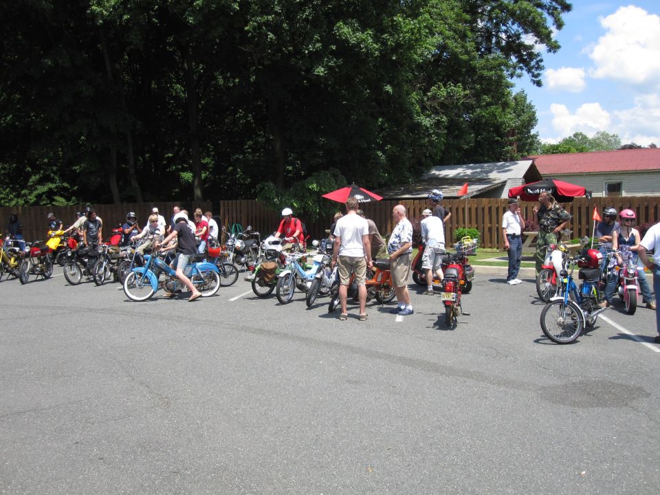 Group of mopeds parked in a lot with people standing around them
