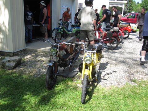 1981 Garelli Rally Sport - Several people gather around two mopeds parked outside a garage on a sunny day.