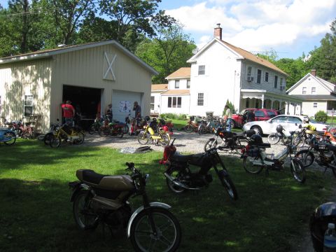 1981 Garelli Rally Sport - A large collection of mopeds and motorcycles are gathered on a lawn in front of a residential property.