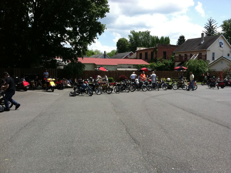 Many mopeds parked in a lot with people standing around