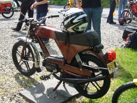 1981 Garelli Rally Sport - A brown moped is parked outdoors with a helmet on its seat, and other vehicles and people are visible in the background.