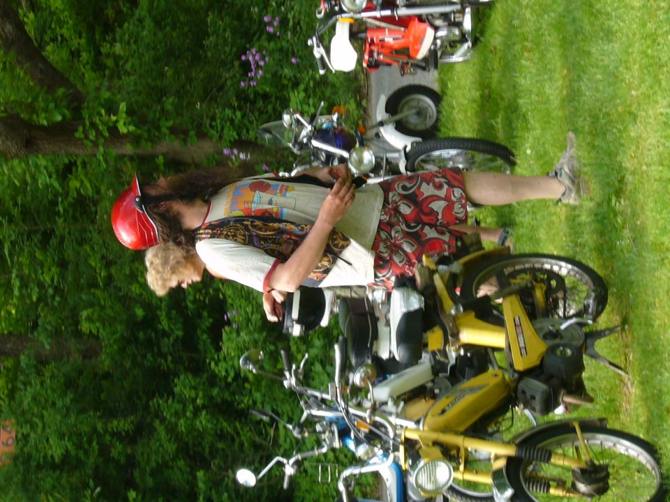 Two people stand near several parked mopeds on a grassy area. One person wears a red helmet and a patterned vest. The other person has blonde hair. A yellow moped is in the foreground