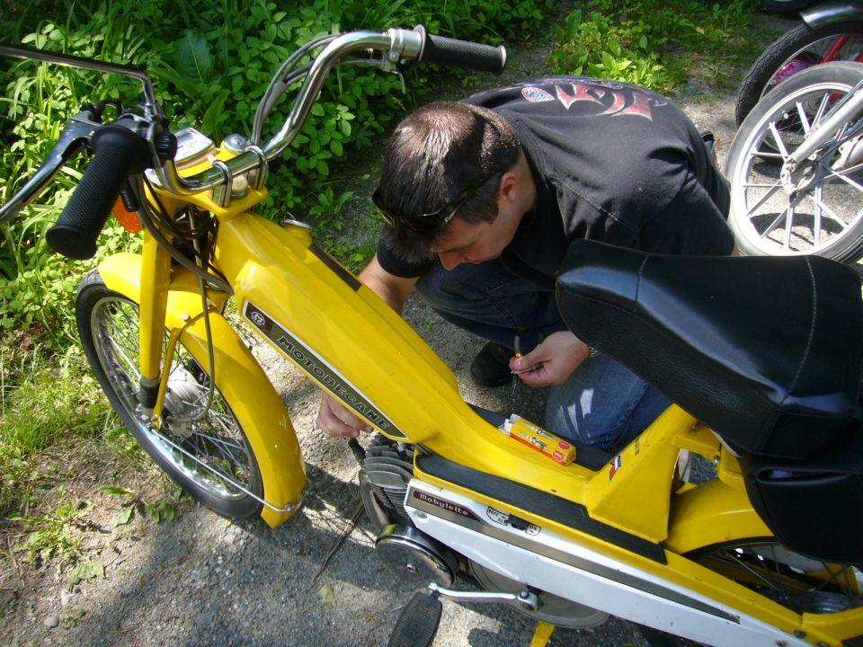 Yellow moped with black seat, chrome handlebars, and a person working on its engine. Another moped wheel is visible