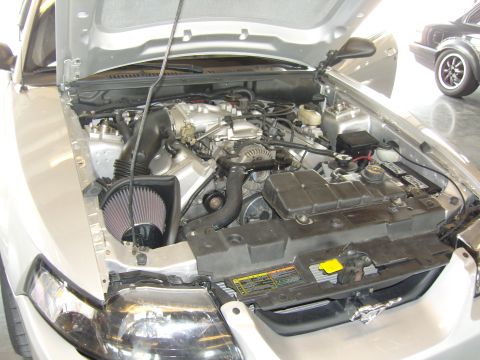 2007 Ford Mustang - The engine bay of a silver Ford Mustang, an aftermarket air intake and other engine components. Another Mustang is visible in the backgroun…