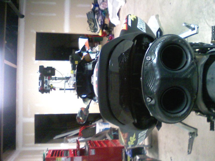 Motorcycle's dual exhaust pipes with carbon fiber accents. Some stickers are visible on the body